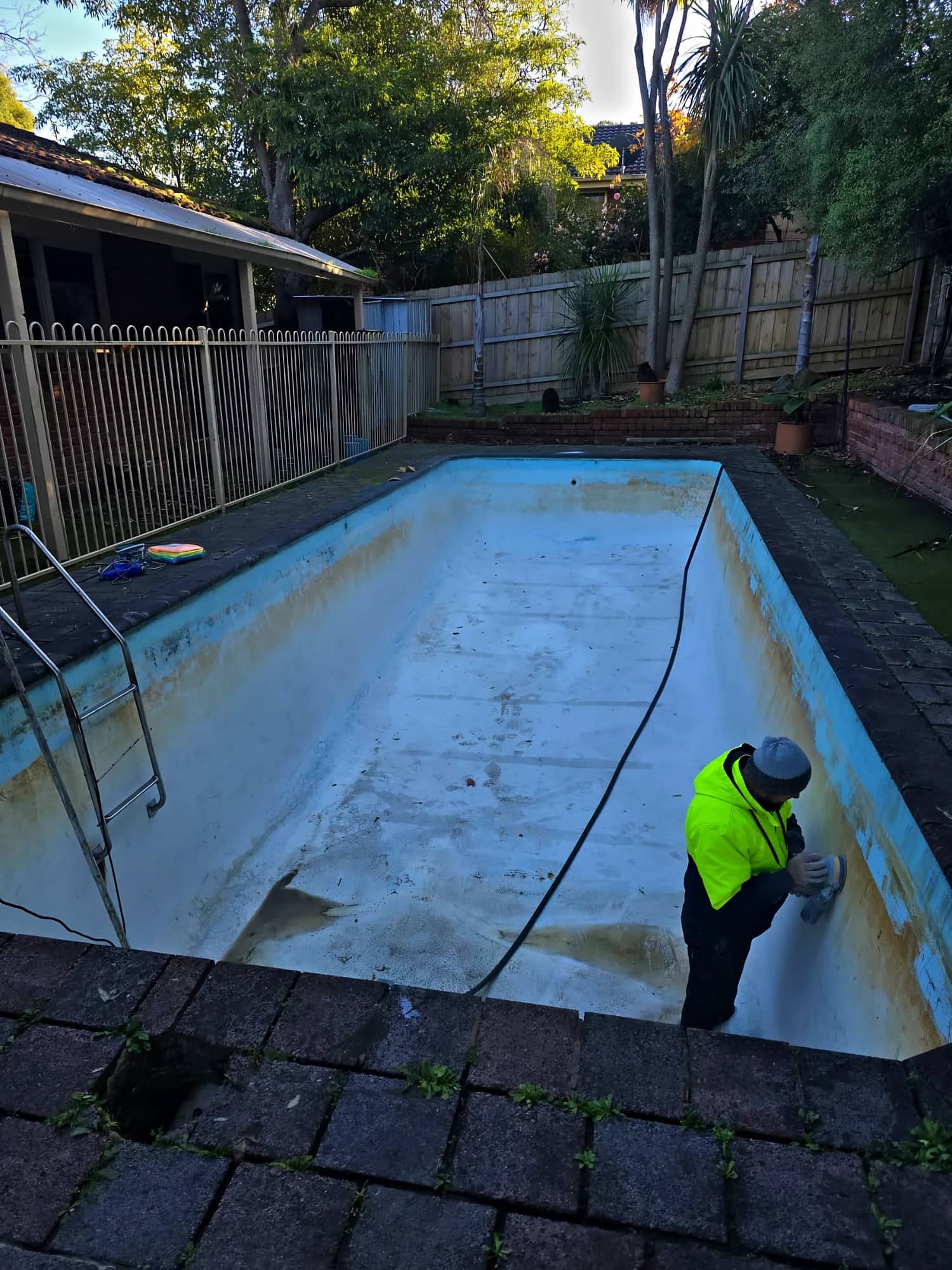 Empty pool shell with a faded, stained surface being prepared for recoating by a worker