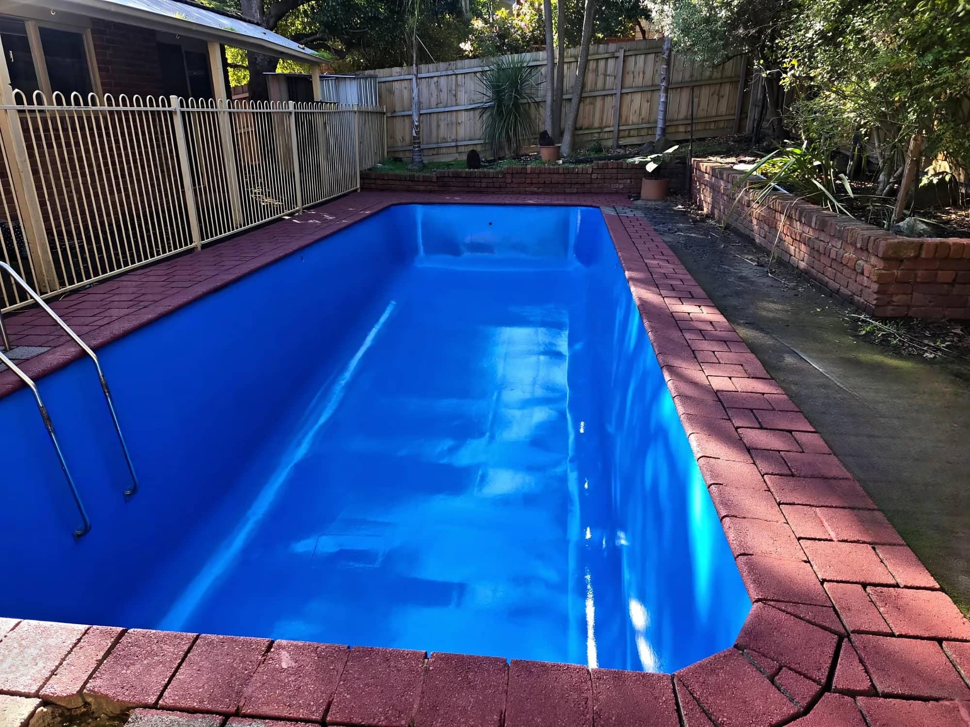 Rectangular pool with a new glossy blue finish, red brick-paver coping and a timber fence