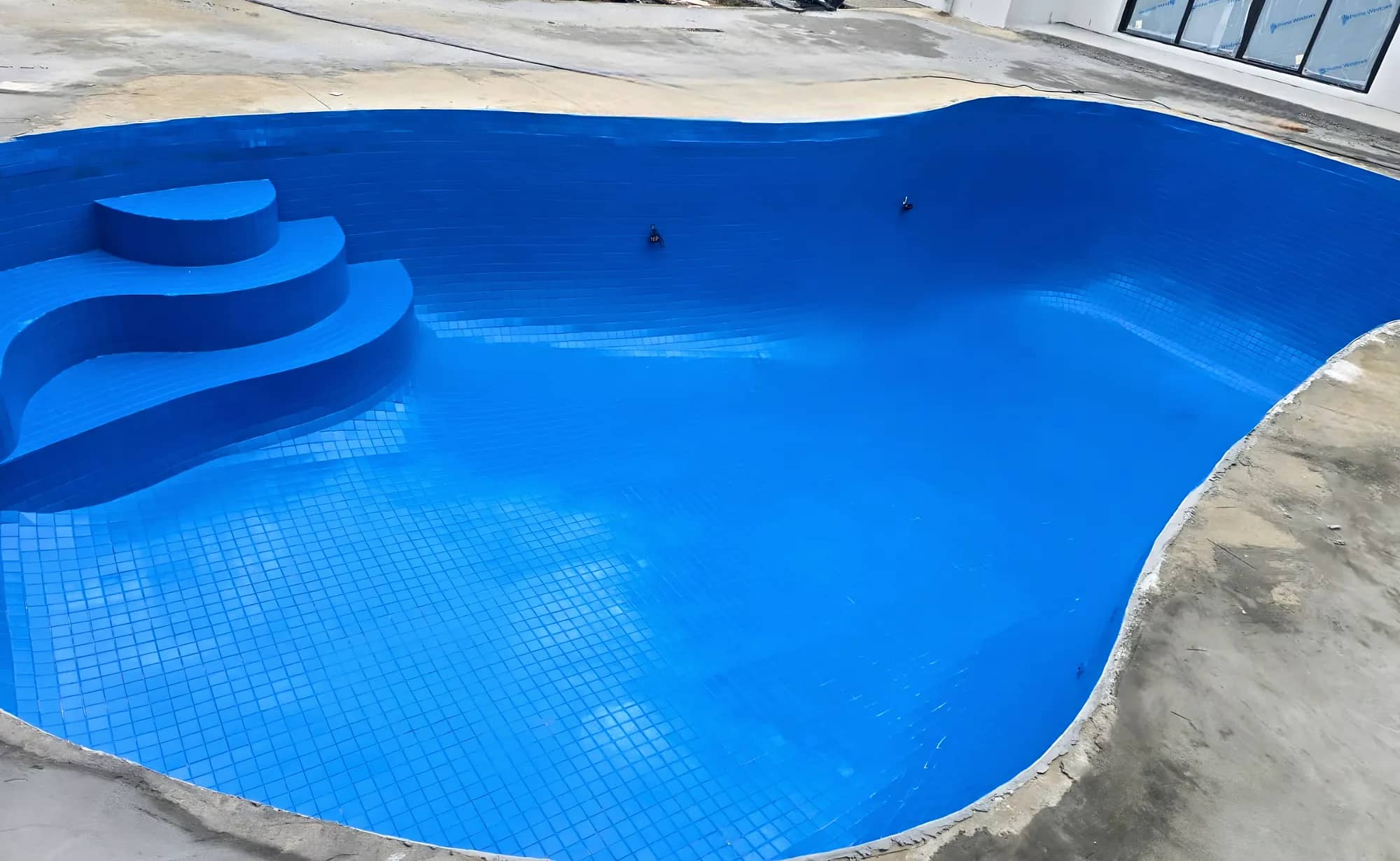 Freeform swimming pool finished in blue mosaic tiles, with wide entry steps, on a new-build site