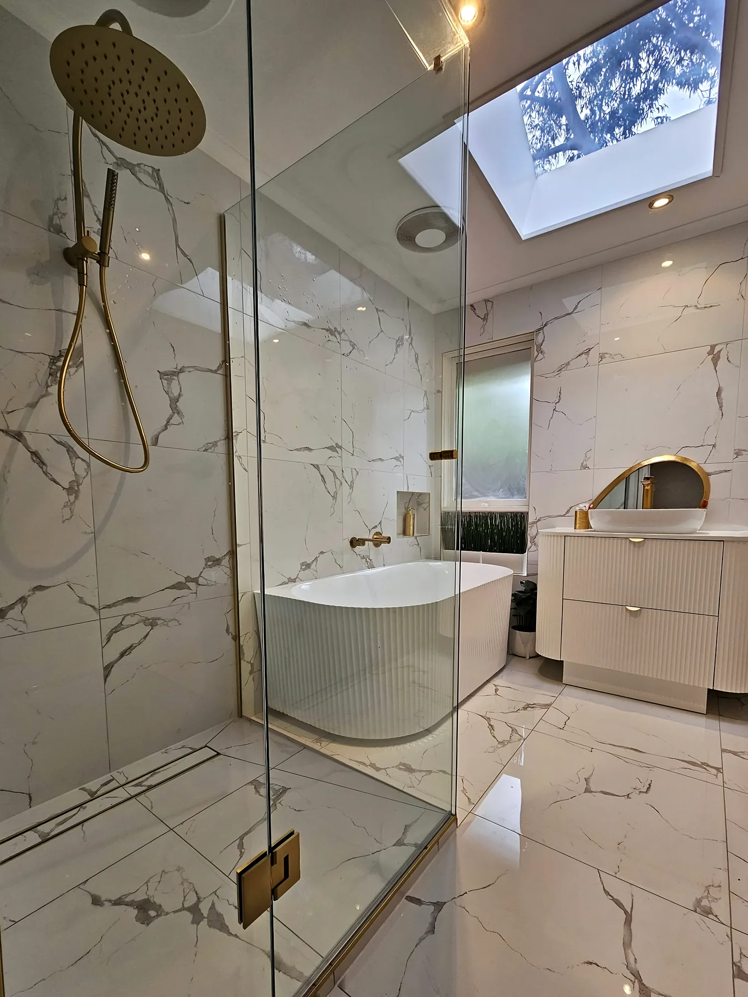 Ensuite bathroom with marble-look wall and floor tiles, a fluted freestanding bath and vanity, brushed-gold tapware and a skylight