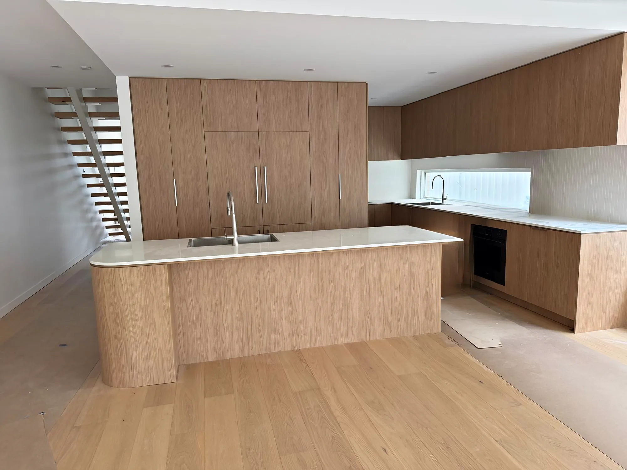 Open-plan kitchen with an oak-look island, tall pantry cabinetry, a white stone benchtop and a floating staircase