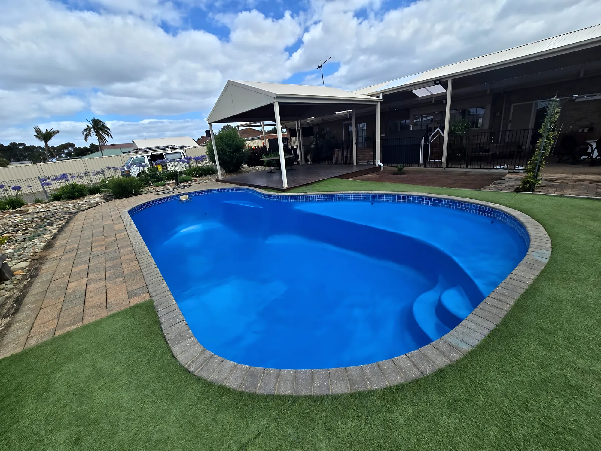 Freshly resurfaced bright-blue kidney-shaped pool with brick-paver coping, synthetic lawn and a pergola under a blue sky
