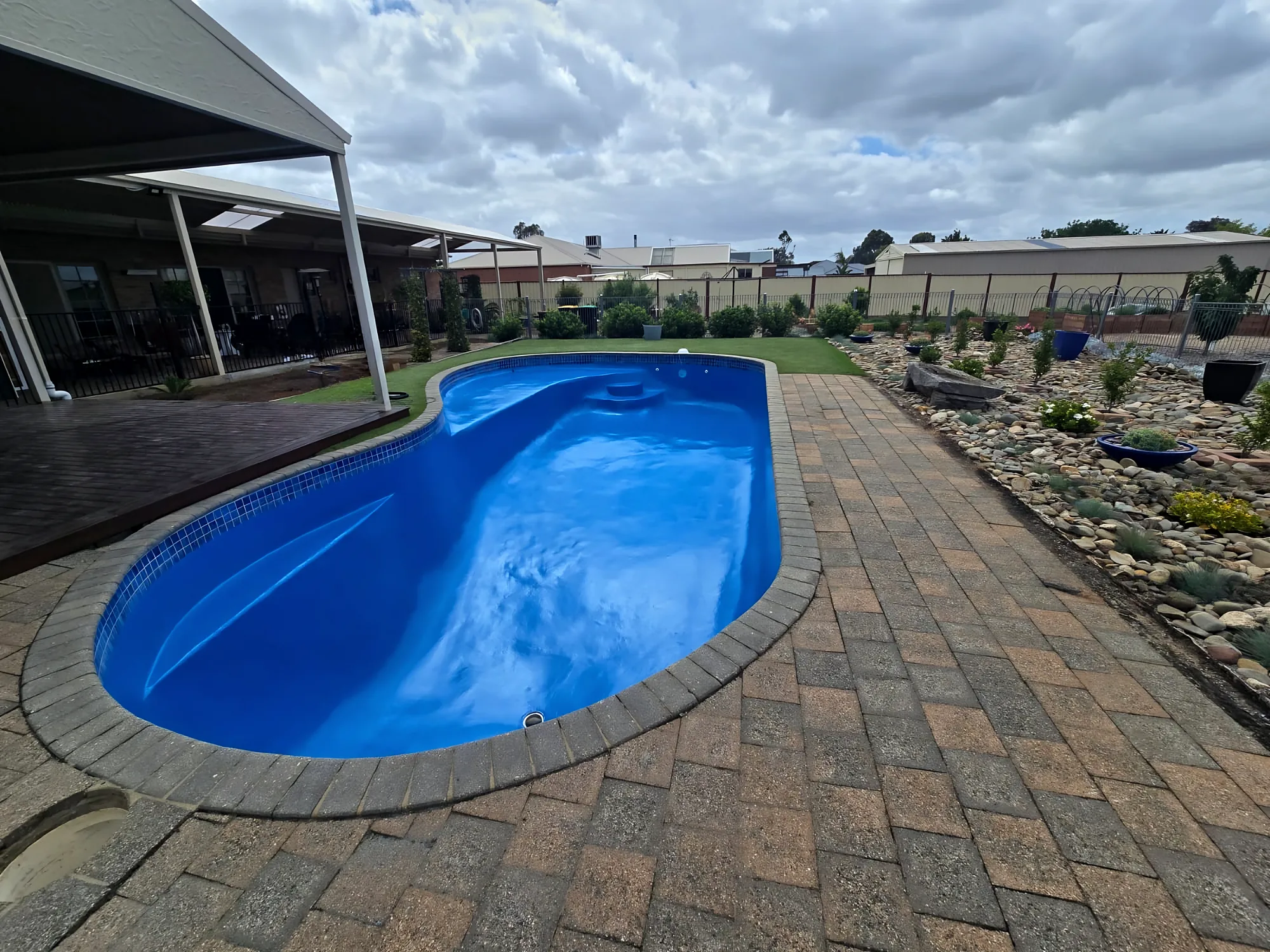 Resurfaced blue kidney-shaped pool seen from the paved side, with garden beds and a cloudy sky