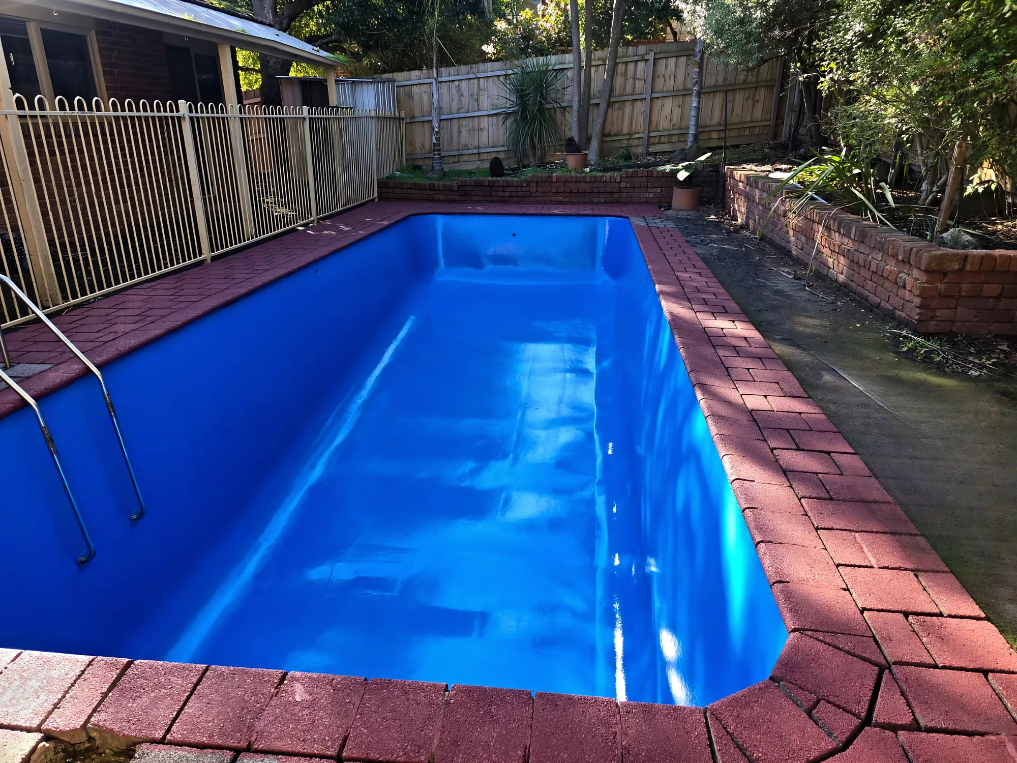 Rectangular pool with a new glossy blue finish, red brick-paver coping and a timber fence