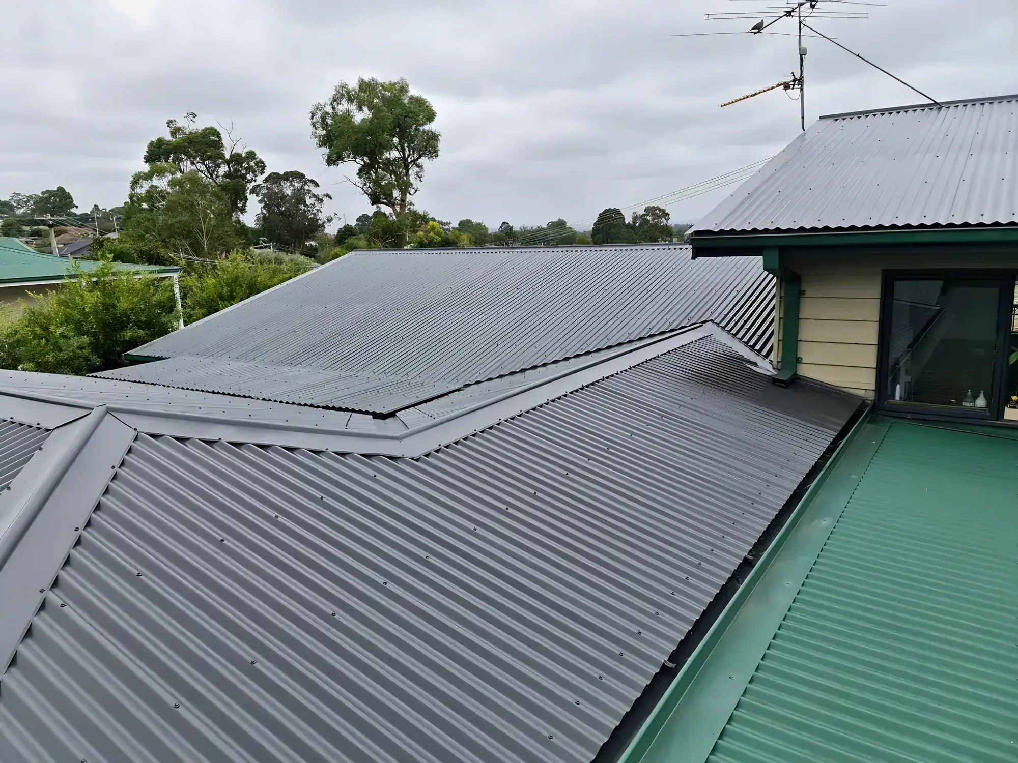 Corrugated metal roof sections finished in grey beside a green metal roof section under a cloudy sky